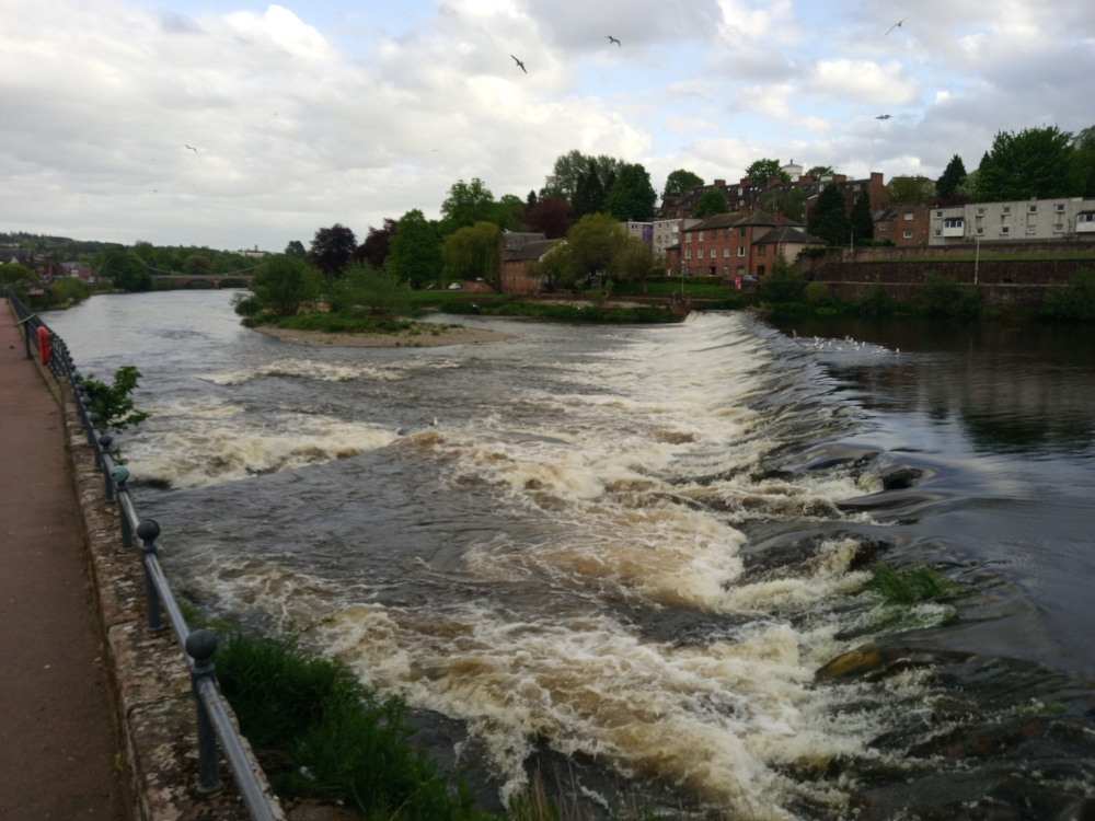 Rushing river, its water a churning mix of dark brown and frothy beige, cascading over a low, stone weir that spans most of the image's width. The water's movement is dynamic, with frothy whitecaps and swirling currents visible throughout. A metal railing, dark grey with rounded finials, runs along the left edge of the image, paralleling a red-brown paved pathway.  Sparse, low-lying vegetation clings to the riverbank near the railing. In the background, a line of brick buildings, a mix of residential houses and what appears to be an apartment complex, rises on a slightly elevated bank. Trees, primarily green and lush with hints of yellow-green, line the riverbank and fill the spaces between the buildings. A few birds are visible in the sky, small against the vastness of the clouds. There's a suggestion of a bridge in the far background spanning the calmer river section before the weir.