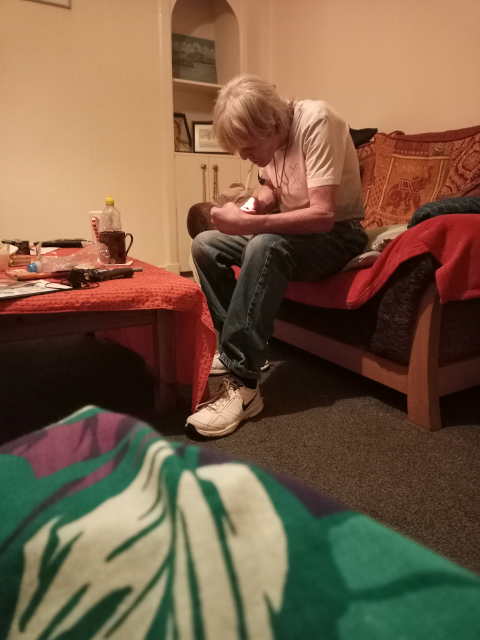 Older man with light, slightly unkempt blond hair. He's seated on a worn, multi-coloured couch, slightly angled to his right. He's wearing a light beige, short-sleeved t-shirt with a barely visible design, and faded blue jeans. His attire suggests casual comfort. He's wearing off-white Nike sneakers. His posture is slightly hunched, and his hands are engaged near his body, seemingly manipulating a small object— possibly a phone or a small device— held close to his chest. His expression is difficult to ascertain due to the angle, but it appears pensive or focused.