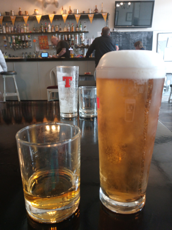 Dark, polished table in a pub setting, showcasing a collection of glassware. Dominating the foreground are: a short, squat tumbler partially filled with a golden amber liquid (likely whiskey or a similar spirit), positioned slightly off-center to the left; and a tall pint glass brimming with a light, golden-hued beer, topped with a thick, frothy head, situated to the right. Between these two, slightly blurred and out of focus, is a smaller, empty glass. Behind them, a taller, frosted pint glass, bearing a red T logo (suggesting a specific brand of beer), sits on the table's surface. In the background, blurred but discernible, are patrons seated at a bar. A man with short, dark hair is visible in the center, wearing a dark jacket, seemingly engrossed in conversation. To his left, another man with a shaved head and a dark shirt is seen from behind, partially obscured, seemingly involved in a task behind the bar. A woman with lighter hair is partially visible to the right, possibly also sitting at the bar.  A muted, darker figure is partly shown in the very far left corner. The setting is a traditional pub with a warmly lit interior. The background shows a long bar stocked with a vast array of liquor bottles neatly organized on shelves. These bottles shimmer under the lighting, creating a sense of depth. Behind the bar, there is a television screen displaying a dark image, adding a layer of subtle contrast. A dark, possibly chalkboard, menu board is partially visible on the back wall. The walls are a light, neutral color, offset by the dark wood of the bar and the darker, polished tabletop. The lighting is softly diffused, casting a warm, inviting ambiance, with subtle highlights reflecting off the surfaces of the glasses.