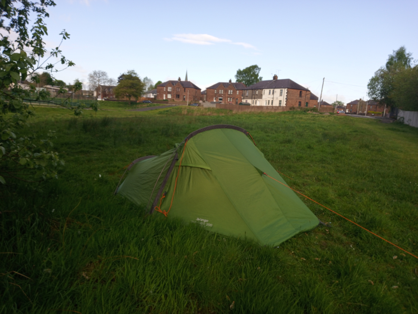 Vibrant, olive-green, single-person dome tent pitched on a grassy field. The tent is partially pitched, with a single guyline visible stretching diagonally from its corner towards the lower right. The tent’s fabric has a subtle sheen, suggesting a lightweight, possibly nylon material. The brand logo, partially visible, indicates a Vango brand. The scene is set in a verdant, slightly overgrown grassy field. The grass is a mix of bright and darker greens, showing some unevenness in height. Behind the tent, a row of modest, two-story brick terraced houses stretches across the background under a pale blue, almost cloudless sky. The houses are muted earth tones—reds and creams—with visible chimneys and somewhat uniform architecture. The lighting suggests late afternoon or early evening, with a soft, diffused light casting few strong shadows.