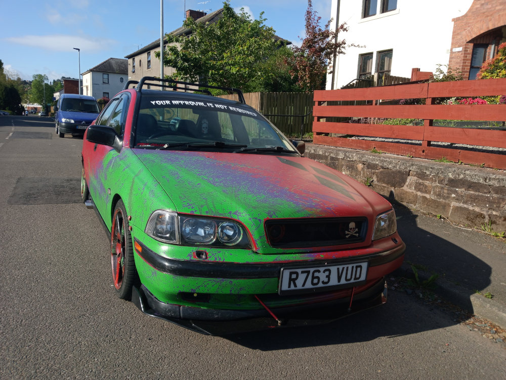 Modified Volvo S40, parked on a residential street. The car is predominantly painted in vibrant, splatter-effect green and red, giving it a chaotic and artistic look. The paint appears deliberately uneven, with drips and splatters suggesting a deliberate, possibly customized, application. The car features a roof rack, a front lip spoiler, and aftermarket wheels. A sticker on the wind shield reads, YOUR APPROVAL IS NOT REQUIRED in a sans-serif font. The license plate reads R763 VUD. A small skull emblem is visible on the front grill. A person is partially visible through the windshield, but only their head and shoulders are seen; they appear to be wearing dark clothing. In the background, a blue van is parked further down the street. There are houses with brick facades and gardens visible. A reddish-brown wooden fence separates the street from the homes' yards.
