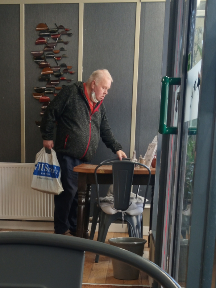 Central figure is an older man with light skin and thinning light-grey hair, slightly receding. He's wearing a dark grey, zip jacket with a red inner lining, dark trousers, and appears to be wearing a light-colored face mask partially obscured under his jacket collar. He holds a white shopping bag with blue lettering (WHSmh) in his left hand and seems to be looking down at something on a nearby table. His posture suggests he's leaning slightly forward, attentive to the table. The table is a rustic, dark-brown wooden table with a somewhat worn appearance. On it are a few indistinct objects that appear to be small containers or condiments, and what looks like a piece of folded paper. A dark grey metal chair with a light grey cushion sits at the table, mostly obscured by the man's body. A grey plastic bucket sits under the chair. The scene is set in what appears to be a cafe or diner. The walls are a muted grey-blue, accented by a wall-mounted metal sculpture composed of numerous dark-red, brown, and bronze-colored leaf-like shapes. A radiator runs along the wall behind the man. The floor is a light-colored wood. A dark-grey door or large window frame dominates the right side of the image, with a green metal handle prominent. There's a hint of greenery and possibly plants visible through the glass panel of the door.