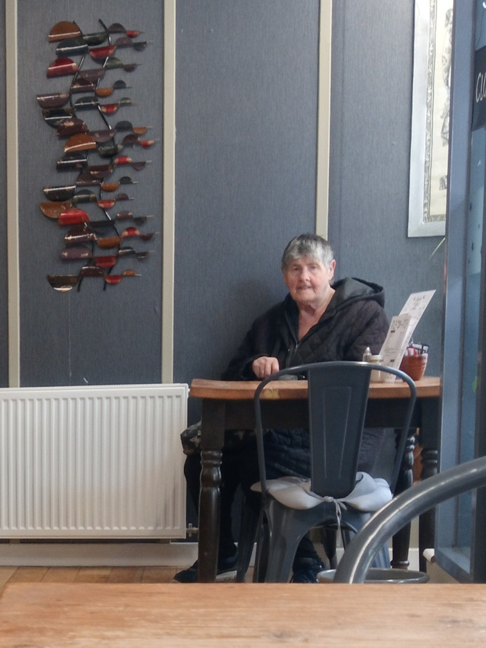 Central figure is an older woman with short, greying hair, seated at a small, square wooden table. Her expression is calm and slightly reserved, almost pensive. She's wearing a dark, possibly black, hooded jacket or coat. Her hands rest loosely on the table. She appears to be alone. A dark gray metal chair with a light gray cushion sits directly in front of her. A portion of another metal chair is visible in the bottom right, suggesting more seating. A white radiator is visible on the wall behind her. On the table, there's a small, partially visible stack of papers or napkins, a small container (possibly sugar or condiments), and a partially visible brown object that could be a small plant pot or bag. A striking wall-mounted piece of art hangs on the wall behind the woman. It consists of numerous small, curved metal pieces in a variety of reddish-brown, dark brown, and muted gold tones, arranged in a cascading pattern that resembles stylized leaves or fish.