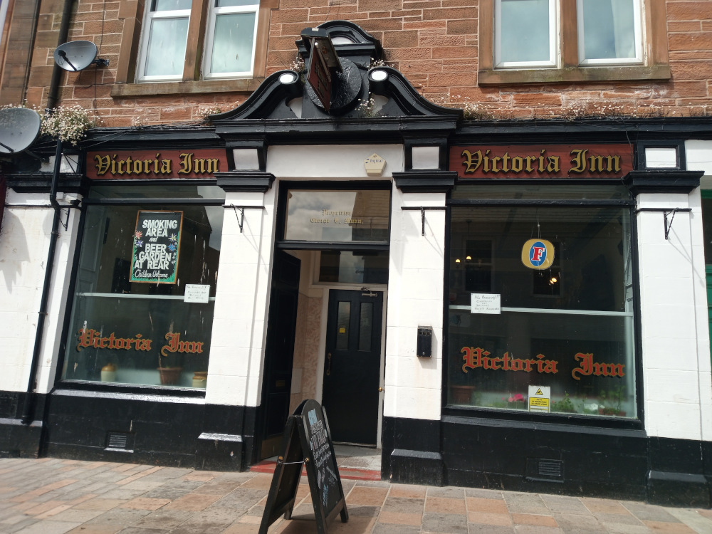 Full shot of the Victoria Inn, a pub seemingly situated on a high street. The pub is a two-story building with a brick façade, a reddish-brown hue subtly variegated by the mortar. The brickwork is clean and shows little weathering. Above the pub’s storefront, the second floor shows two windows, suggesting residential space above the commercial area. The pub itself is characterised by a prominent, dark-coloured, ornate cornice and fascia above the store front. The building's sign, Victoria Inn, is crafted in elegant, gold-coloured lettering on a deep reddish-brown panel, which is framed within a black, decorative moulding. The photograph is taken from a slightly low angle, directed toward the center of the pub's façade. The composition is straightforward, focusing directly on the building with some sidewalk visible at the bottom. The symmetrical nature of the building is emphasised by this composition, which directs the viewer's attention to the pub's entryway and central design elements.