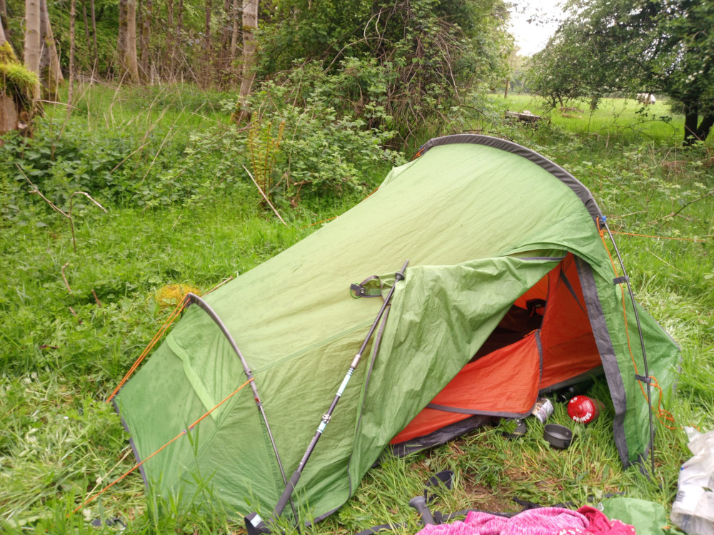 Central focus is a vibrant green and orange one-person backpacking tent, pitched haphazardly in a grassy area. It's slightly ajar, revealing an orange interior. A pair of trekking poles leans against the tent's side, their teal and black colours contrasting with the tent's brighter hues. Inside the tent, glimpses of a small, reddish-orange sleeping bag or liner are visible. Near the tent's entrance, a small red canister, possibly a gas canister, and a dark cooking pot rest on the grass. At the bottom right, a portion of a pink or red item, potentially a piece of clothing, is partially visible. The overall condition of the tent suggests it has been used. The scene is set in a verdant woodland. Lush green grass, speckled with wildflowers and interspersed with low-lying shrubs and trees, forms the immediate foreground and background. The trees are a mix of deciduous species, with their leaves fully developed, suggesting spring or summer. The lighting suggests an overcast day; the light is soft and diffused, with no harsh shadows.