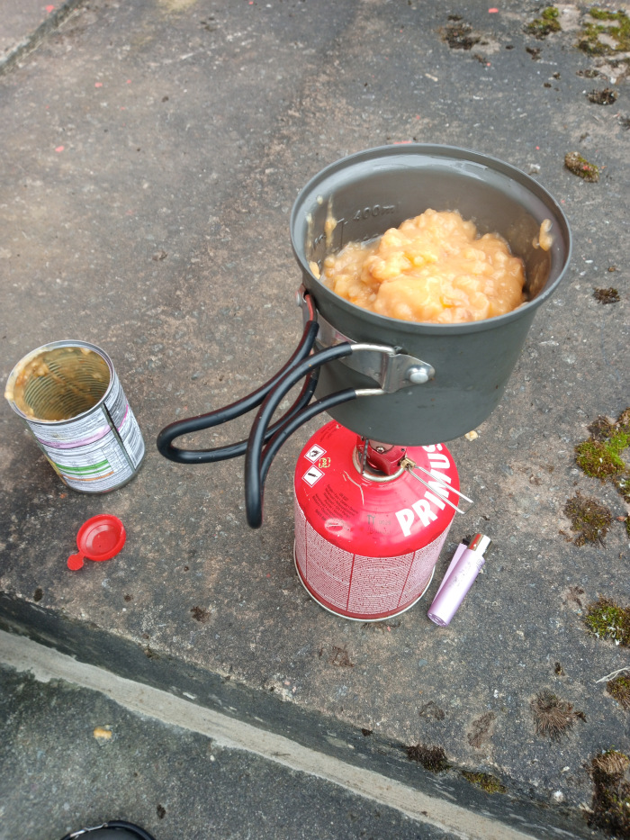 Grey camping pot perched atop a red Primus camping stove. The pot is about two-thirds full of a pale, creamy-orange substance, possibly some sort of cooked fruit or pudding, that appears thick and chunky. The pot has a slightly metallic sheen and a simple design. The stove is cylindrical, red with white text and safety warnings. A black metal stand supports the pot over the stove. To the left, an empty, slightly crumpled tin can with some residual pale orange contents rests on its side. The label is partially visible, indicating it may contain food. A small, red plastic cap lies nearby, likely from the can.
