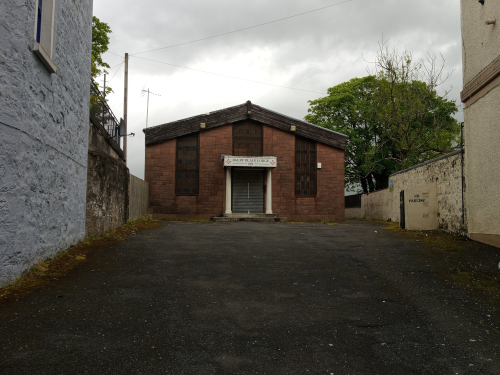 Single-story, rectangular building of reddish-brown brick. Its gable roof is steeply pitched, and the building's facade is austere. A simple, rectangular sign reading DALRY BLAIR LODGE is affixed to the building's upper centre. The entrance is a dark, closed metal roll-up door. Flanking the building on both sides are aged, light-grey and dark grey textured walls forming a narrow alleyway. The walls show signs of weathering and age, with patches of moss and discoloration. A faded NO PARKING sign is visible on the right-hand wall. Trees with full, green foliage provide a backdrop to the scene.