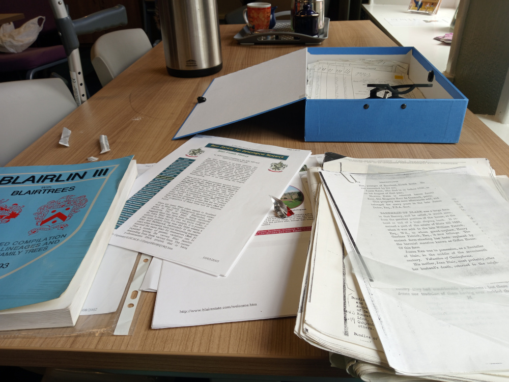 Light brown wooden table cluttered with genealogical research materials. Dominating the scene are stacks of papers, some neatly organised in folders, others haphazardly piled. A large, pale blue, lidded box sits slightly to the upper right, possibly containing more documents. A thick, blue book titled BLAIRLIN III with BLAIRTREES lies prominently on the left, its spine clearly visible. Other printed material includes what appears to be a smaller booklet or pamphlet with a crest on it, and several loose sheets of paper, some handwritten, some typed. A metallic object, possibly a small paperweight or letter opener, rests near the center. In the upper left corner, a portion of a chair and a cane are visible, hinting at a domestic or care setting. A thermal flask sits partially visible behind the papers towards the top of the image.