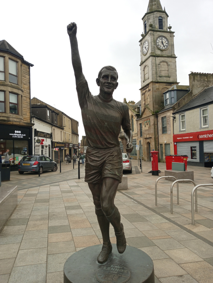 Bronze statue of a dynamic male figure, seemingly a footballer, captured mid-stride with his right arm raised in a triumphant gesture. The statue is realistically rendered, showing detailed musculature and the texture of clothing. He is wearing what appears to be a short-sleeved, striped athletic shirt and shorts, and calf-high socks with boots. His facial expression is one of exhilaration, his mouth slightly open, and his gaze directed forward and slightly upward. The statue stands on a circular dark-grey granite or stone pedestal, upon which a small, barely visible plaque is affixed. Around the statue, the background includes pedestrians (mostly blurred) moving casually on a paved town square, their clothes exhibiting a range of muted earth tones and blacks. There are buildings lining the street.