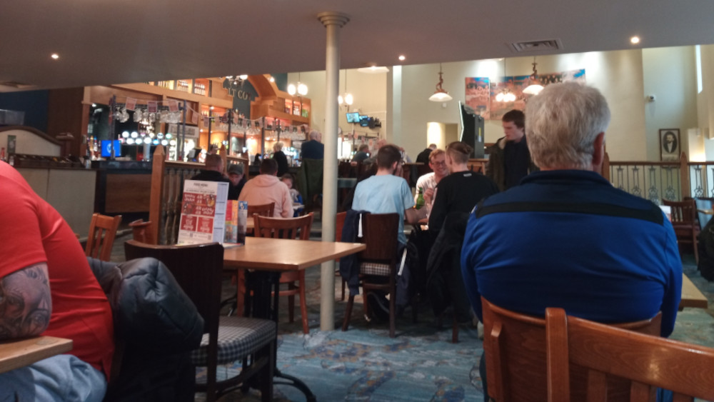 Bustling pub interior, viewed from a slightly low perspective, as if seated amongst the patrons. The focal point is a group of people seated at tables, engaged in conversation or eating.