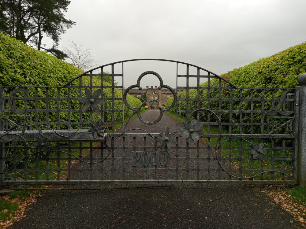 Central focus is an ornate, wrought-iron gate, positioned slightly off-centre, dominating the foreground. The gate is dark gray-green, appearing aged and possibly weathered, with a slightly rustic finish. It features a central, arched top section, embellished with a decorative quatrefoil (four-leaf clover) motif. The gate’s lower portion displays a grid-like structure, interspersed with stylised, three-dimensional flower designs sculpted into the metal. The number 2000 is prominently displayed near the base. The gate is closed.