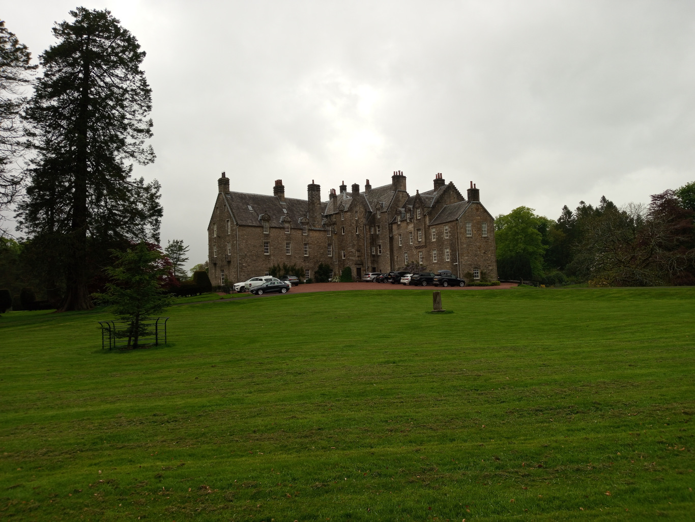Central focus is a large, imposing stone mansion, typical of a Scottish country house or castle. It's a multi-story structure with multiple gables, numerous windows, and a grey-brown stone exterior. Several cars are parked in a line in front of the building, suggesting a driveway or parking area. A small, dark grey car is prominently positioned slightly off-center, just in front of the mansion.  Other cars are less distinct. In the foreground, a lush, vibrant green lawn stretches across the entire lower half of the image. A simple, black metal garden structure, possibly a small trellis or plant stand, stands alone on the left side of the lawn, mid-ground. A lone, relatively small deciduous tree stands near the garden structure, creating contrast in scale and texture with the tall conifer in the upper left.
