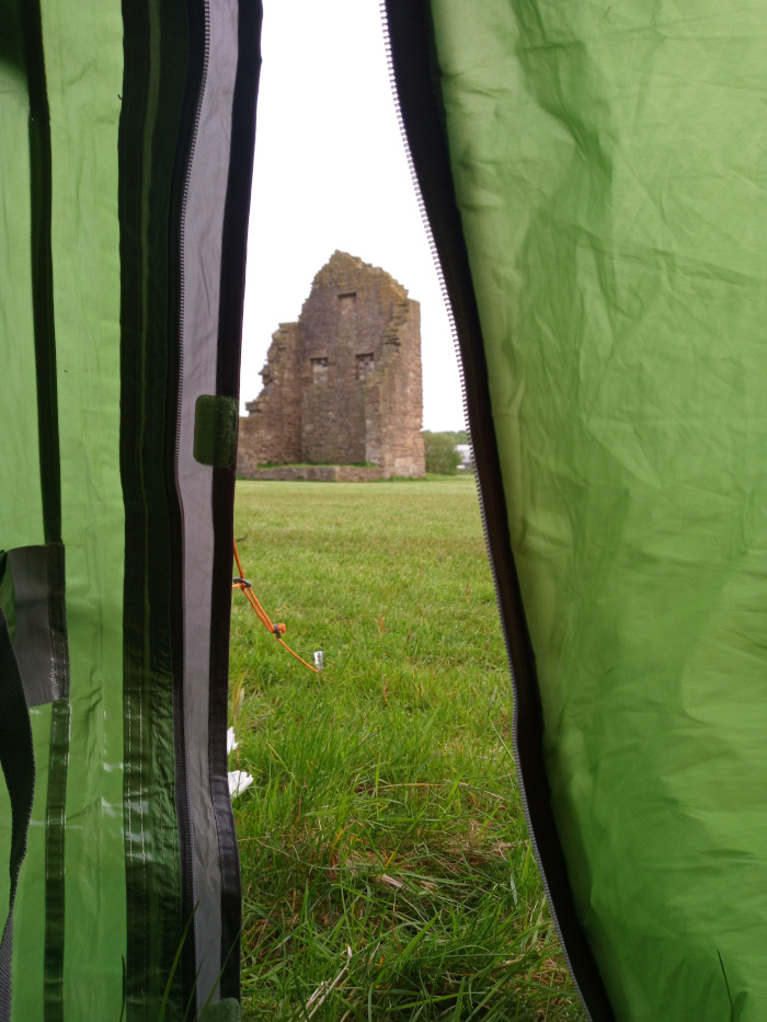 Predominantly a view from inside a green tent, specifically from between two slightly ajar sections of the tent's fabric. The tent's material is a vibrant, slightly crumpled, lime-green nylon-like fabric with darker grey zipper seams running vertically. A small, light grey plastic piece is visible near the upper-left side of the grey seam. An orange-brown cord is partially visible, extending from inside the tent towards the outside. The main focus is on the view through the tent opening, which reveals a stone ruin in the mid-ground. The ruin is a substantial, grey stone structure with visible holes that suggest windows or former openings. It's in a state of disrepair, with portions visibly missing and showing its age.  The background beyond the ruin consists of a field of tall, lush, vibrant green grass. No people are visible in the image.