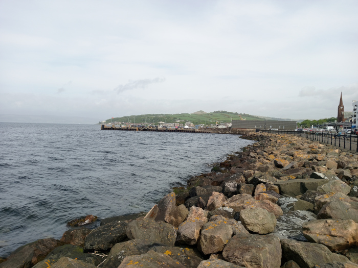 Tranquil coastal scene, dominated by a rocky shoreline gently sloping into a calm, dark-blue sea. The immediate foreground is a chaotic yet aesthetically pleasing arrangement of large, irregularly shaped rocks. These rocks vary in color from dark grays and browns to patches of ochre and lichen-covered surfaces, suggesting age and exposure to the elements. The rocks are damp, with hints of subtle water flow visible in crevices. No people are present in the foreground. Beyond the breakwater, a small, charming town nestles against a gently sloping hill, its buildings a mix of muted colors—mostly browns, greys, and whites. A tall, slender church steeple, a reddish-brown hue, is a prominent vertical element in the right background. The hill itself is covered with a blanket of verdant green vegetation, hinting at a temperate climate.
