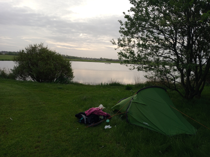 Vibrant green, single-person tent pitched on a grassy bank. It's slightly angled, its entrance facing away from the viewer toward a calm body of water. Near the tent, a pile of camping gear is visible, including a dark-colored backpack and a purplish-pink item that appears to be clothing. Some crumpled white trash or tissue is scattered near the gear. There is no human figure directly visible, but a pile of clothing near the tent suggests a person has recently been there. The scene unfolds at the edge of a tranquil lake or pond. The water is relatively still, reflecting a muted sky. The far bank of the water is low and mostly flat, with a line of trees and shrubs creating a soft, horizon line.  The sky above is overcast, with soft, diffused light that lacks harsh shadows.