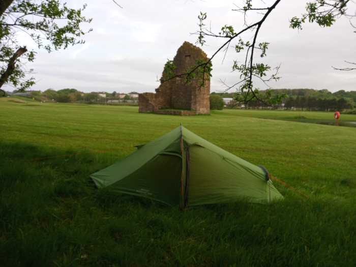 Vibrant, olive-green, single-person backpacking tent pitched in a lush, verdant field. The tent is positioned slightly off-centre, angled to the left, with its entrance partially obscured. Its fabric appears taut and wrinkle-free, suggesting a recent setup. Thin orange guy lines are subtly visible, extending from the tent's corners into the grass. In the background, a significant portion of a crumbling stone ruin, possibly an old castle or tower, stands prominently against the horizon. The ruin is predominantly gray-brown, with patches of missing stone revealing the sky. Its form is irregular and aged, showing clear signs of deterioration.