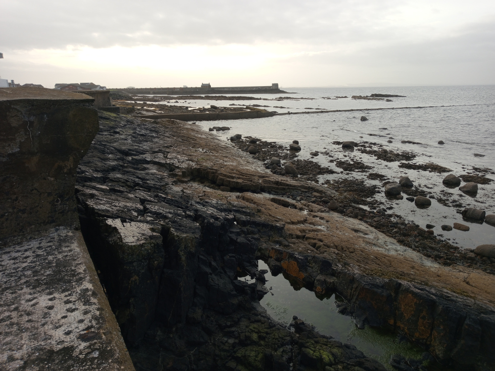 Sark, rocky coastline scene. There are no people present. The main focus is on a dark, fractured rock formation extending from the foreground diagonally toward the middle ground. This rock is primarily dark brown and black, showing signs of weathering and erosion with visible fissures and crevices. Patches of lighter brown and orange are visible within the darker rock, possibly due to mineral deposits or differing rock strata. A small, still pool of dark, greenish water sits nestled within a deeper crevice in the foreground rocks. In the background, a low, grey concrete structure, possibly a seawall or remnants of a building, runs horizontally across the image. Beyond this, further out to sea, there are other low-lying structures, appearing somewhat distant and indistinct, possibly buildings or other coastal installations. Scattered boulders of various sizes are visible both in the foreground, along the shoreline, and further out in the shallow water. The sea itself is relatively calm, a dark greyish-blue, reflecting the muted light of the sky.