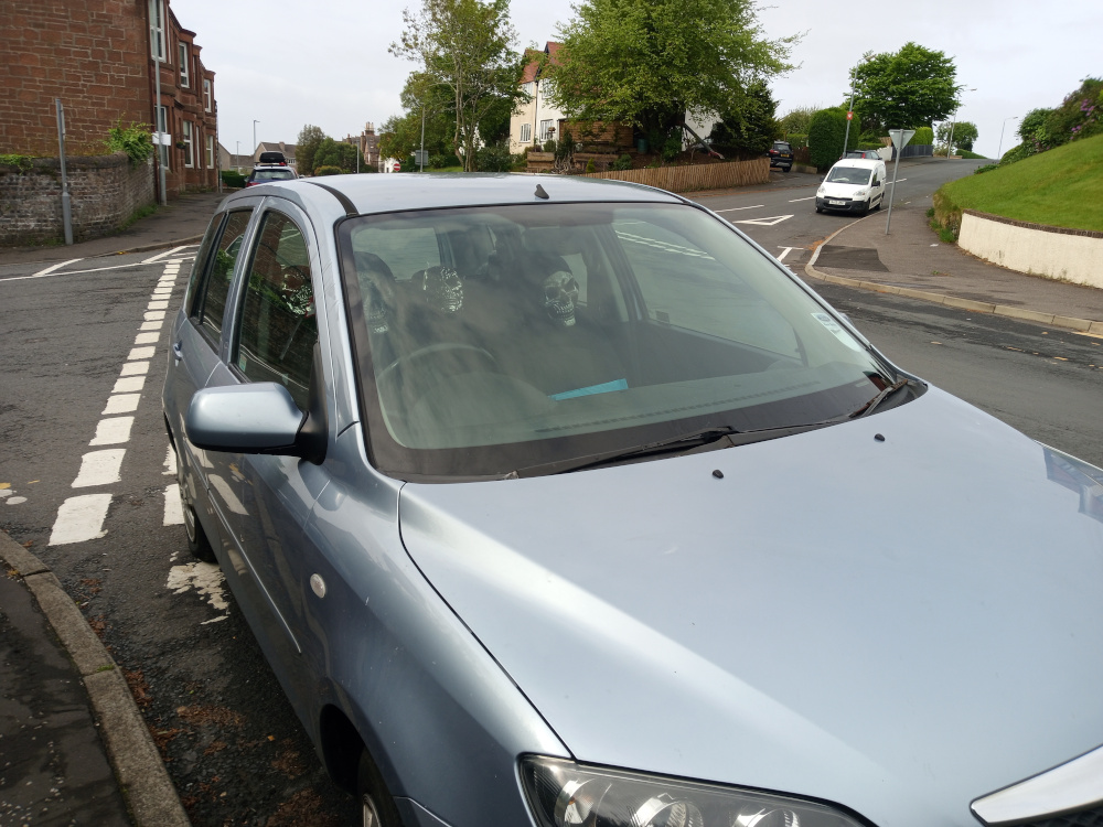 Light blue Mazda 2 hatchback parked on a residential street. The car is angled slightly to the left, showcasing its driver's side. Inside, what appear to be two skull-shaped objects are visible on the dashboard. The driver and any potential passengers are not clearly visible, though their presence is implied. In the background, a white van is parked further down the street, and another smaller vehicle is barely visible behind the Mazda. The scene is set on a typical residential street, with a mix of red brick and stone buildings in the background. The street is relatively quiet with some greenery and trees visible along the sides. The day is overcast, the lighting is soft and diffused, creating even illumination without harsh shadows.