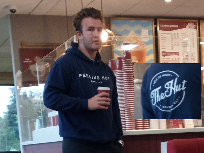 Young, light-skinned man with short, dark brown hair styled upwards. He has a strong jawline and a neutral, almost pensive expression. He's wearing a navy blue hooded sweatshirt with the words FEELING HUT SINCE '05 in white lettering across the chest. His sweatshirt is slightly oversized, with the sleeves pushed slightly up his forearms. He's holding a Costa Coffee takeaway cup in his right hand, the cup's red and white design partially visible. He stands slightly angled to his left, his body occupying most of the foreground. He appears to be in a relaxed yet slightly withdrawn posture. Below his sweatshirt, a hint of dark-coloured pants is visible. In the lower right corner, a portion of another navy blue sweatshirt is shown, this one displaying a circular logo with the words The Hut, Isle of Wight, Colwyn Bay in a stylised script font. This sweatshirt is out of focus and appears to be part of a different person, possibly not fully in the frame.