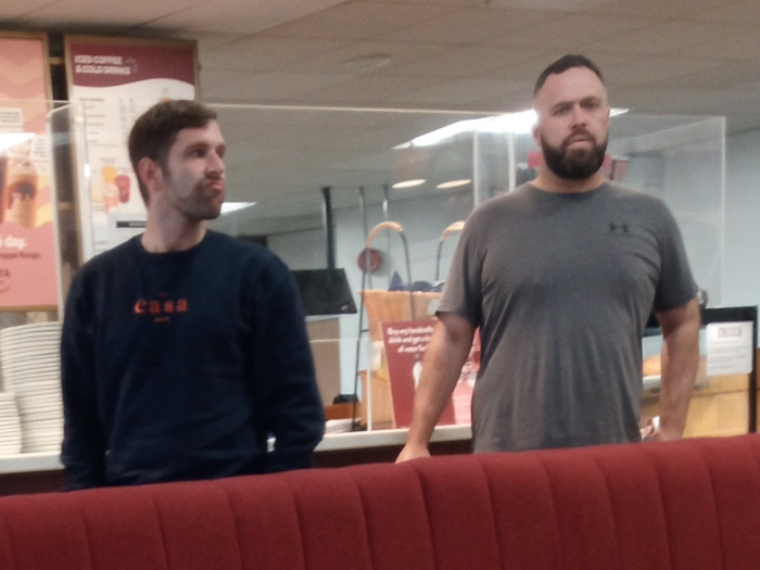 Two men in what appears to be a casual dining establishment. First man has short, dark brown hair, a slightly stubbled beard, and is wearing a dark navy blue crew neck sweatshirt with the word Casa in a reddish-orange script across the chest. His expression is somewhat neutral, perhaps slightly pensive or bored, and his gaze is directed slightly to the right, away from the viewer. His body is positioned relatively straight, his hands are likely resting in his lap (obscured by the booth). Second man is larger and has short, dark hair cut very close to his scalp. He has a fuller, well-groomed beard and mustache, darker than his hair. He's wearing a gray Under Armour branded t-shirt. His posture is relaxed but upright, his arms at his sides. His expression is serious and possibly slightly stern. He appears to be looking directly forward. Behind the men, a partially visible menu board featuring drink options, primarily in shades of beige and brown, is prominent. A clear plastic partition or screen stands between the men and a partially visible counter area. Some light brown paper bags are visible on the counter. The overall background is a muted blend of off-white walls and various shades of light gray and beige.
