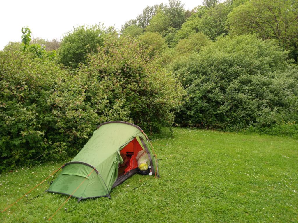 Vibrant green, single-person tent, pitched on a lush, slightly damp-looking lawn. The tent is a dome shape with orange interior peeking from the open doorway, suggesting it's partially packed or ready for occupancy. Orange guy lines are subtly visible, extending from the tent stakes to anchor it. Inside the tent, a hint of orange and yellow suggests sleeping bag or other gear. The tent sits slightly angled, not perfectly parallel to the viewer's perspective.