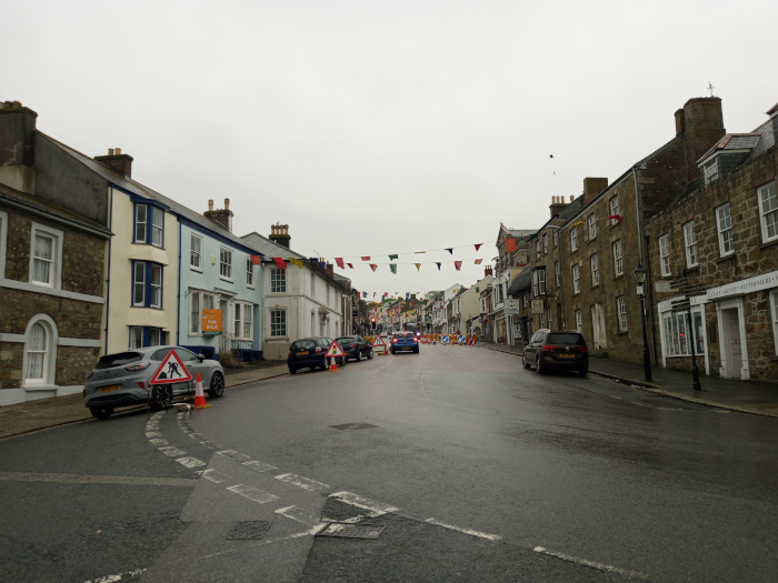 Street scene in what appears to be a small, historic town. The main focus is a street, wet from recent rain, lined with two-story stone and brick buildings. Several cars are parked along the sides of the street and a few are driving down the road. One light grey Ford Puma is parked prominently on the left, near the foreground, with traffic cones beside it, suggesting roadworks. A dark brown SUV is visible further down the street, parked on the right. Other vehicles are scattered throughout the scene but are less distinct. There are no people visible in the image.