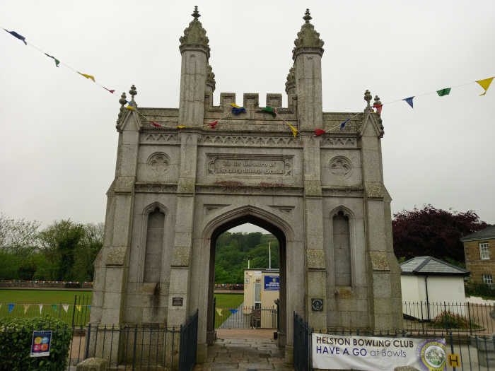Dominant feature, taking up most of the frame. It's a substantial stone structure, resembling a miniature castle gatehouse. Two tall, slender towers flank a central arch, topped with crenellations (battlements). Detailed stone carvings are visible on the archway's facade, including what appears to be an inscription near the top. The stone is a light grey, showing signs of weathering and age. The arch is open, revealing a pathway and a glimpse of greenery beyond. A vibrant, rectangular sign is affixed to the base of the archway's lower portion. It boldly proclaims HELSTON BOWLING CLUB HAVE A GO at Bowls in clear, sans-serif lettering, contrasting the muted tones of the stone. The sign incorporates the club's logo and is brightly coloured, likely using a palette of blues, yellows, and perhaps greens.
