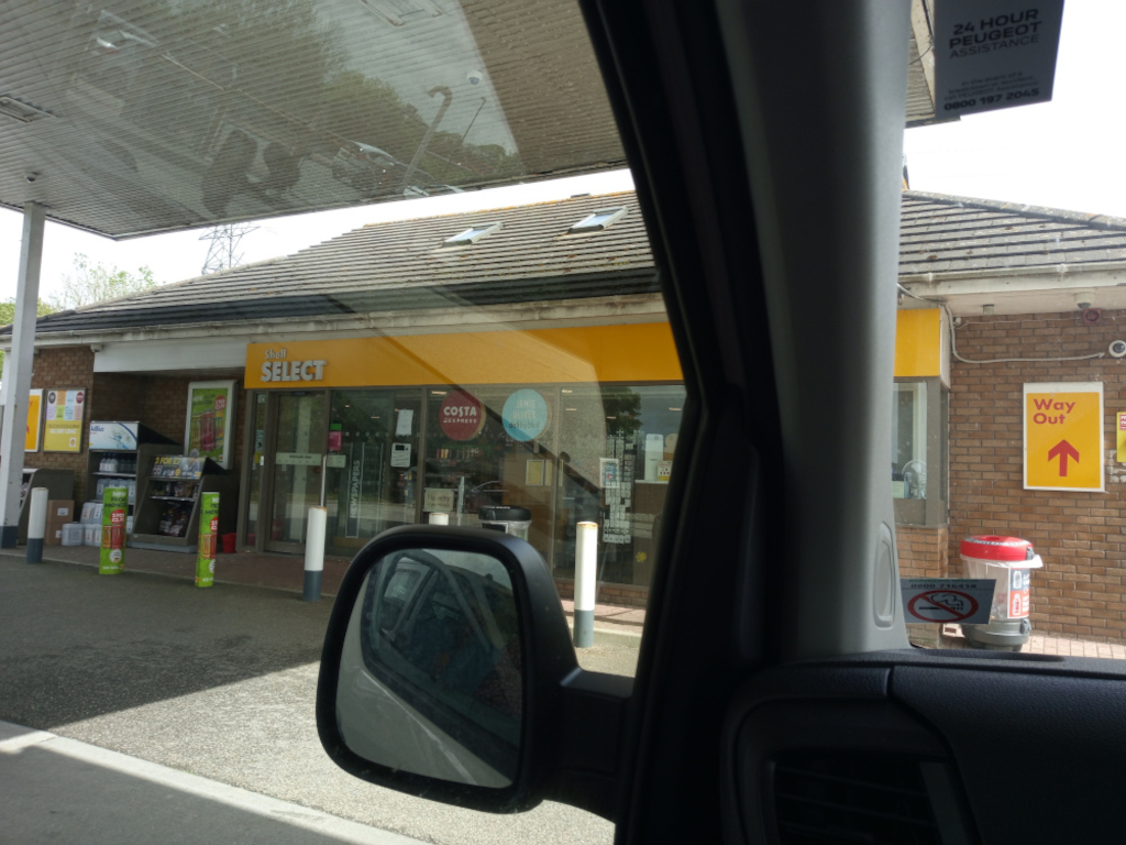 Shell Select convenience store situated at a petrol station. The store's facade is predominantly yellow, with a prominent Shell SELECT logo in bold lettering. A Costa Coffee logo is visible within the store's window. Inside, various goods are displayed on shelves; newspapers are subtly suggested. The store has large glass windows that showcase the interior. Outside, there are white bollards lining the store's entrance, and a Way Out sign with an upward-pointing arrow is on the brick wall of the building. A red cylindrical bin is partially visible near the exit. A car's interior, specifically the driver's side, dominates the foreground, creating a frame-within-a-frame effect. The car's side mirror reflects a distorted view of the scene. No people are directly visible.