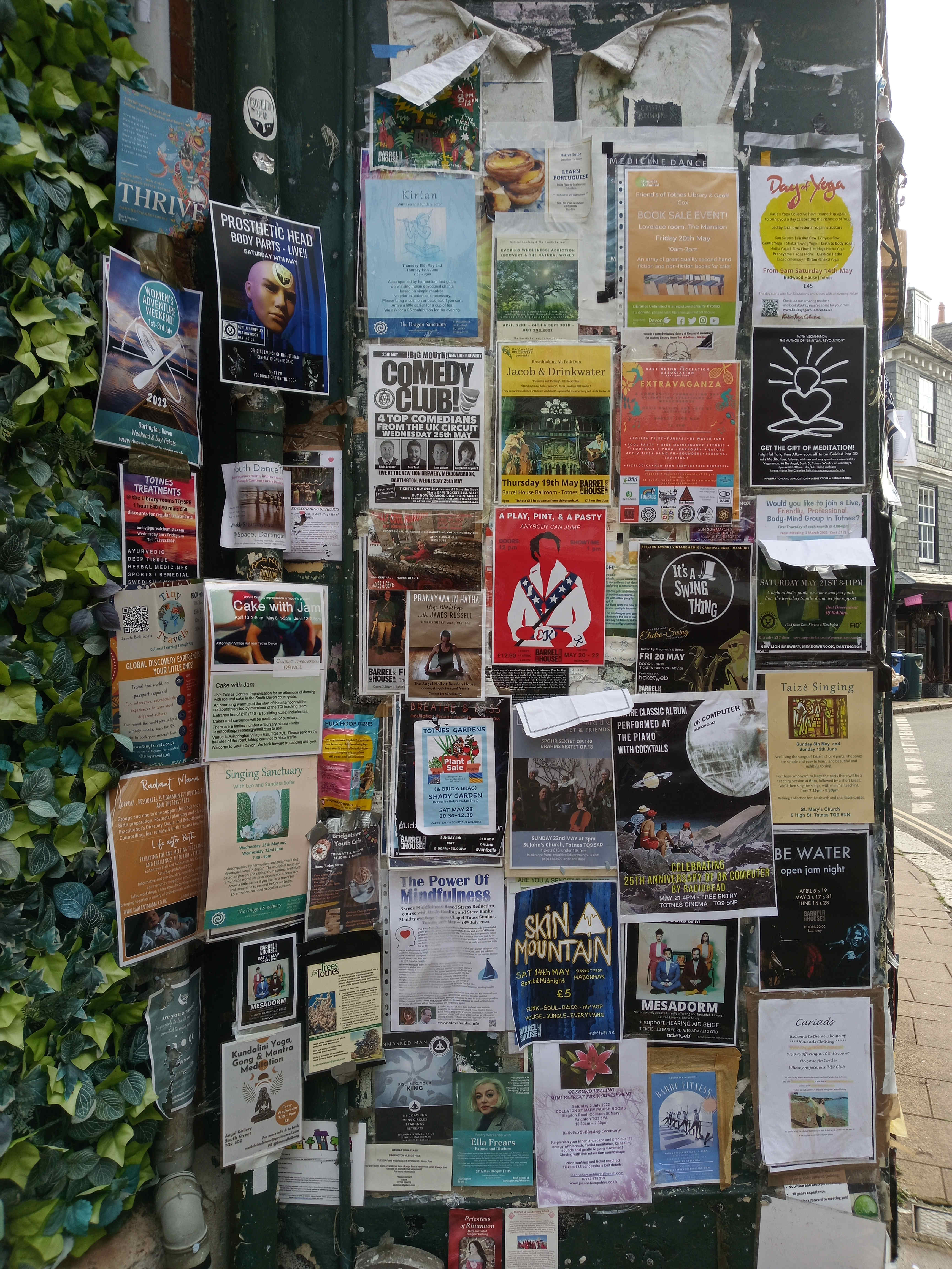 Dark green, slightly weathered notice board overflowing with a chaotic tapestry of flyers and posters. These advertisements are densely packed, overlapping and vying for attention. The flyers advertise a diverse range of events and services: yoga classes, comedy shows, musical performances (including a Radiohead tribute), book sales, workshops (meditation, mindfulness, dance), community events, and even a plant sale. The posters are varied in size, color, and design style, ranging from minimalist and text-heavy to illustrative and image-rich. Some are neatly affixed, while others appear more haphazardly stuck, with corners peeling and edges creased. There are no people directly visible on the board; instead, the posters feature photographs or illustrations of performers, instructors, or product imagery.