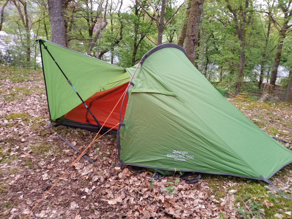 Green Vango Banshee 200 tent, pitched in a woodland setting. The tent is a single-wall design, with a distinctive, slightly curved peak. Its main body is a deep olive green, showing subtle creases and folds indicating its fabric texture. A bright orange inner tent is partially visible through the entrance. Attached to the tent is a lime-green footprint or awning extending outwards, supported by a dark grey pole. Orange guy lines, taut and slender, radiate from various points on the tent, anchoring it to the ground. The groundsheet is visible under the tent, dark grey in color. A small, dark object, possibly a backpack, is partially concealed beneath the tent near its base. The scene is set within a deciduous forest. Tall, slender trees with lush green canopies create a dappled light effect on the forest floor. The ground is covered with a thick layer of dried brown and reddish-brown leaves, creating a textured, earthy surface. The overall palette is muted and natural, featuring earthy browns, deep greens, and the contrasting bright orange and lime green of the tent. 