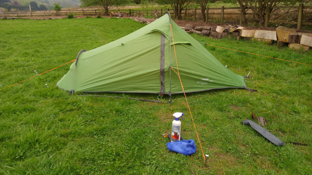Bright, lime-green, one-person backpacking tent, pitched on a verdant lawn. It's a single-layer dome tent with orange guy lines extending from its corners, anchored into the grass with pegs. The tent is slightly angled, not perfectly symmetrical. A dark grey tent pole is visible running vertically along the centre. The tent's brand, seemingly Vango, and potentially a model name, are subtly printed on its side. Beside the tent, on the ground, are: A spray bottle of what looks like tent cleaner or waterproofing solution. The label is partially visible, with text and a green image suggestive of outdoor use. A bright blue microfibre cleaning cloth, slightly crumpled. A dark grey, narrow, rolled-up item lying horizontally on the grass, possibly a tent pole or a stake bag.