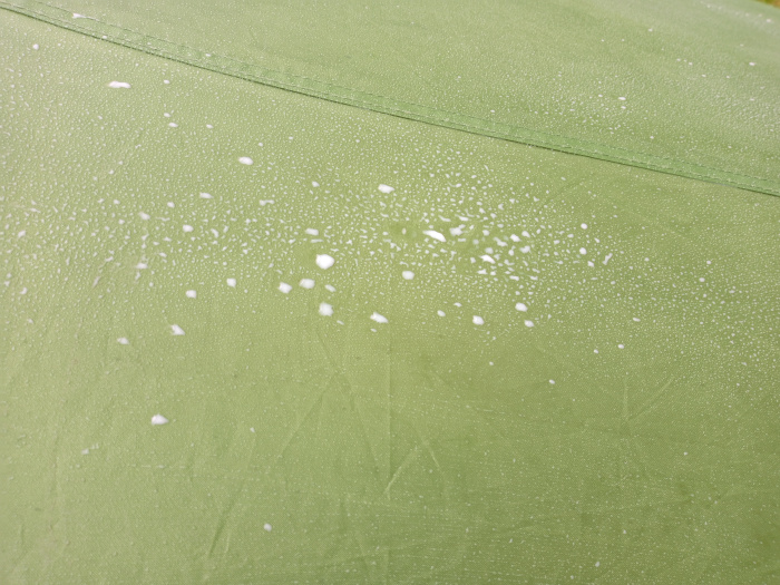 Close-up, slightly high-angle view of a section of olive-green fabric, possibly a tent or awning material. The main focus is the fabric itself, which exhibits a slightly textured, matte surface. Scattered across the fabric are numerous small, irregular white droplets or specks, resembling soap suds or condensation. These droplets are not uniformly distributed, clustering more densely in some areas and sparsely in others. There's a subtle seam or stitching line running diagonally across the frame, creating a subtle division in the surface. No objects or people are present.