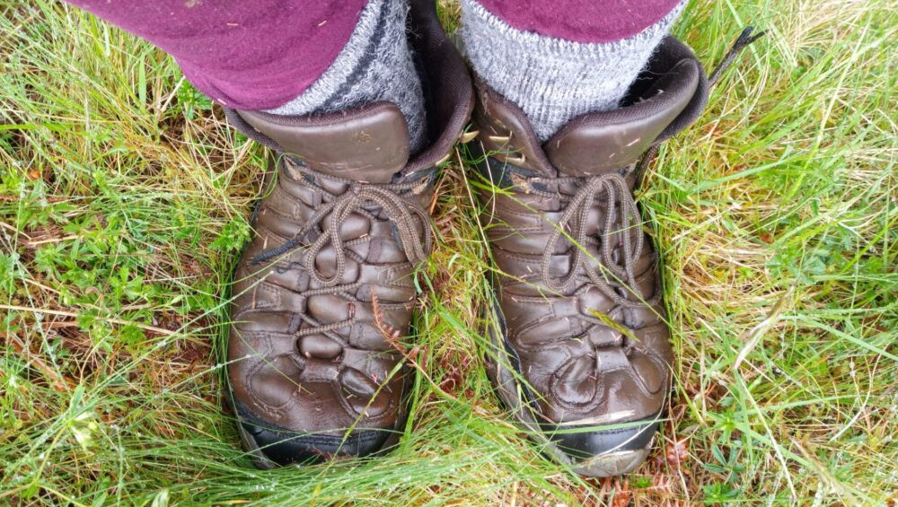 Close-up, slightly high-angle shot centred on a pair of worn, brown leather hiking boots. The boots are laced up and appear damp, showing signs of use and exposure to the elements. They are positioned in the center of the frame, slightly angled outwards. The boots are being worn by a person whose lower legs and feet are visible. We see dark maroon or burgundy leggings and grey wool hiking socks pulled up to just below the calf. The person is stationary, their legs slightly apart.