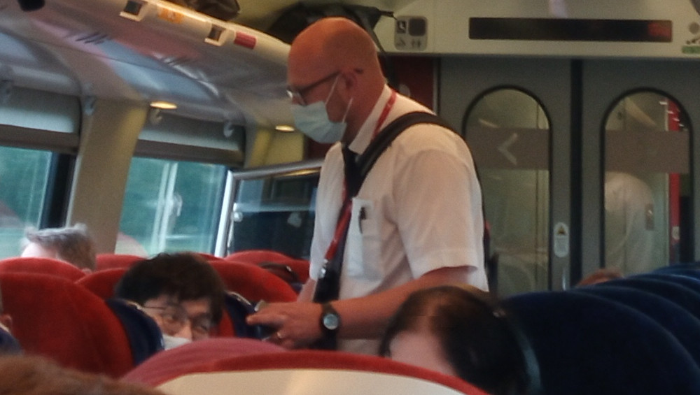 Bald, light-skinned man in his 40s or 50s, wearing a crisp white, long-sleeved shirt, a dark tie, and a dark-red lanyard around his neck. He also sports a light-blue surgical-style mask covering his nose and mouth, and a dark-colored wristwatch on his left wrist. He appears to be a train attendant or conductor, given his attire and the setting. He's standing in the aisle of a train, slightly turned toward the viewer, his body angled towards the back of the train. He's subtly holding something in his left hand, possibly a ticket or device, while his right hand is slightly lower. Surrounding him are several passengers seated in red train seats. One passenger, in the foreground, is a person with dark hair whose face is partially obscured and slightly tilted downward; it looks like they might be sleeping or resting. Other passengers are partially visible, their faces mostly indistinct.