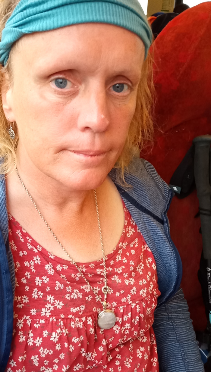Close-up portrait of a fair-skinned woman (Leonie) with shoulder-length, slightly wavy light-brown hair. She's positioned slightly off-centre, facing the viewer directly. She appears to be seated, as suggested by the background. Her expression is neutral, perhaps slightly pensive or tired, with a soft focus in her light blue eyes. She wears a teal ribbed headband that pushes her hair back, a red top with a delicate white floral pattern, and a denim-toned jacket or sweater. A simple silver chain necklace with a small, round pendant hangs around her neck. Small silver earrings are visible in her ears. Her eyebrows are light and relatively thin. Her skin shows some sun exposure or freckles. The background is blurred but reveals part of a red fabric seat (possibly a bus or train seat) and a portion of what seems to be a dark grey or black trekking pole, partially visible on the right. The setting appears to be indoors, within a vehicle of some kind, given the red seat and pole. The lighting is soft and diffused, seemingly natural light possibly coming from a window outside the vehicle, lacking harsh shadows. The overall color palette is warm, dominated by the reds of the clothing and seat, contrasted with the cool teal of the headband and the muted blues and greys of the jacket and background elements.