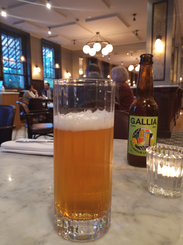 Tall, clear glass of light amber beer, slightly foamy at the top. The glass has a subtly textured, possibly ribbed, design. Beside it, slightly behind and to the right, sits a bottle of Gallia beer, dark amber in colour, with a distinctly stylised label featuring a yellow-green graphic design and text. A small, clear glass candleholder with a lit candle rests near the beer bottle. A crisp white napkin and silverware are subtly visible on the marble table. Blurred but discernible figures sit at tables in the background, suggesting a bustling restaurant or pub setting. The people are dressed in casual attire, mostly muted tones. Their forms are indistinct, serving primarily to suggest depth and context.