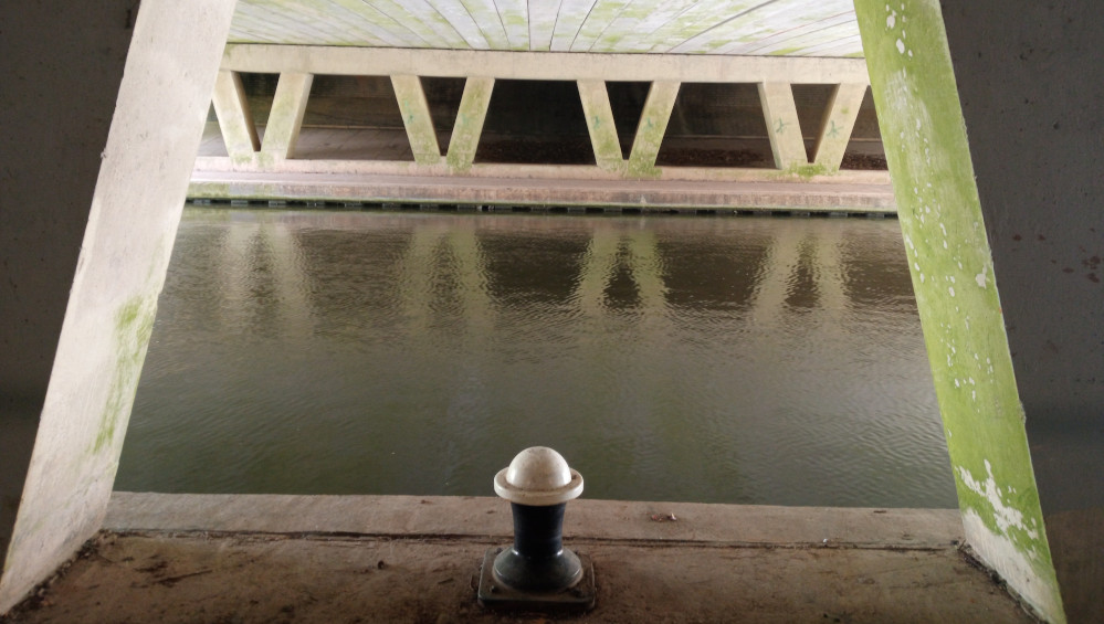 Tranquil canal seen through a triangular opening formed by the supporting concrete structures of a bridge. The bridge's underside is light beige concrete, showing significant age and discoloration, with patches of pale green algae or lichen visible, particularly on the vertical support beams. The canal's water is a calm, murky olive-green, reflecting the muted light and the bridge's structure above. The walls of the bridge framing the view are also concrete, showing similar age and discoloration as the under-bridge structure. The overall lighting is soft and diffuse, suggesting either an overcast day or subdued natural light. The colours are muted and earthy: beige, olive green, dark grey, and light brown.