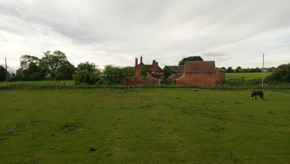 Verdant pasture, occupying the majority of the foreground and extending to a low fence. A lone dark horse, predominantly black with possibly a small white marking (difficult to discern fully), grazes peacefully near the center-right of the pasture. Its posture is relaxed, suggesting a tranquil moment. Behind the fence sits a cluster of brick buildings, predominantly a two-story, gabled house with chimneys and a slightly lower, attached barn-like structure. These buildings are rendered in muted brick reds and oranges, showing some age and weathering. The architecture appears traditional, possibly rural English. In the distance, behind the buildings, are lines of low-lying structures and trees, indicative of a typical English countryside village.