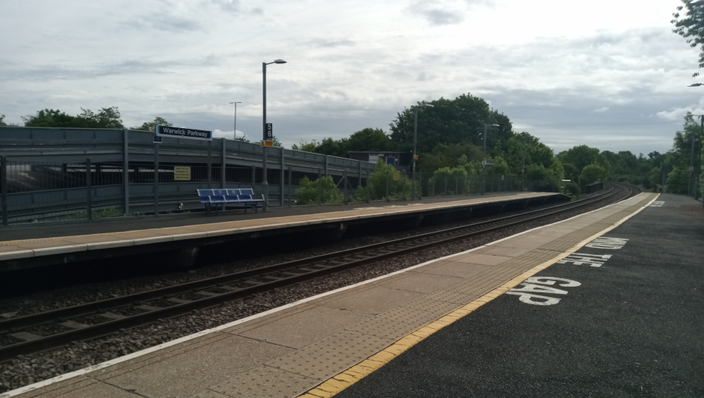 Nearly deserted train platform at Warwick Parkway station. The platform is long and stretches into the distance, disappearing around a gentle curve. It features a light grey concrete surface with a tactile paving strip for visually impaired passengers. A bright yellow line delineates the edge of the platform, and the words Mind the Gap are stencilled in white on the dark asphalt next to it. Three blue metal benches sit unoccupied near the station building. The foreground is dominated by the platform and tracks, while the background includes the station's car park structure, which is grey metal with a multi-level design. There are no people present.