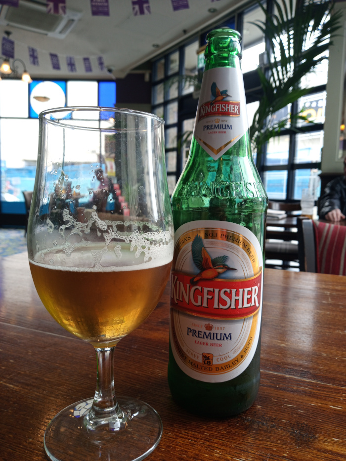 ull bottle of Kingfisher Premium Lager beer dominates the lower-middle portion of the image. It's a dark green glass bottle, reflecting some light. The label is predominantly red and white, featuring the Kingfisher logo (a bird in flight) and text highlighting PREMIUM LAGER BEER and SINCE 1857. The bottle stands upright on a dark wooden table. Blurred figures are visible in the background through the pub's windows and inside the establishment, suggesting a lively yet indistinct atmosphere. One partially visible person in the far background appears to be seated at a table.