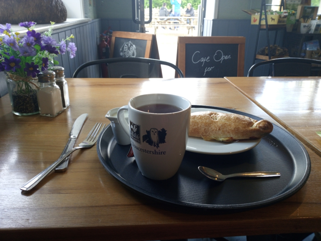 Simple café scene. A wooden table holds a black tray with a mug of tea, a pastry (possibly a Cornish pasty), a small spoon, and cutlery. Salt and pepper shakers and a small vase of purple flowers are also visible at the edge of the table. In the background, a cafe sign says Cafe Open for... and a partially visible outdoor seating area is visible through a window. The overall mood is peaceful and suggests a relaxing break.