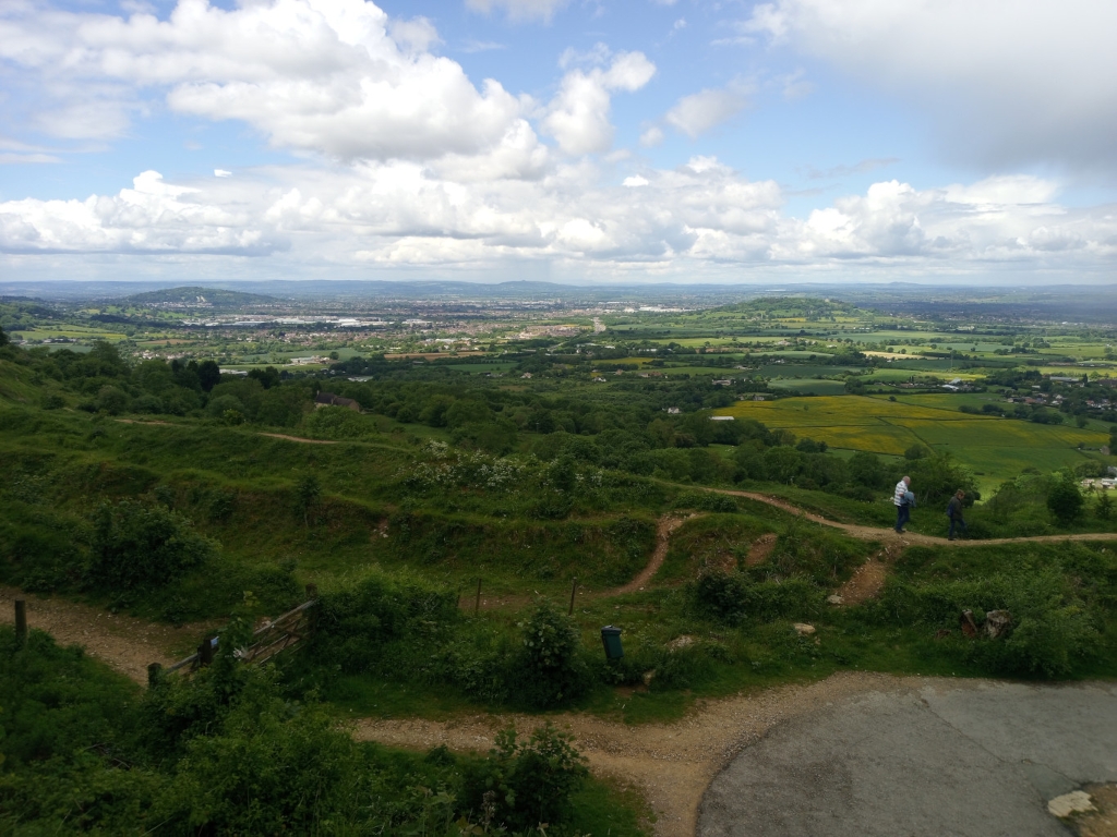 Panoramic view of a sprawling countryside landscape, taken from an elevated vantage point. Rolling green hills dotted with patches of yellow flowers dominate the foreground, leading the eye to a distant cityscape visible on the horizon. Two individuals are seen walking along a dirt path that winds through the hills. The sky is partly cloudy, adding depth and texture to the scene. The overall impression is one of vastness and the beauty of the natural world contrasted with human settlement in the distance.