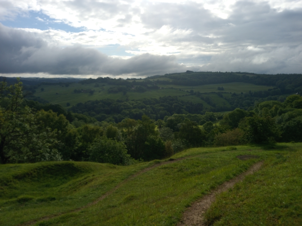 Panoramic view of a verdant landscape under a partly cloudy sky.  The foreground is a gently sloping grassy hill with faint paths. The mid-ground and background consist of rolling hills covered in lush green forests and fields, extending to the horizon. The overall atmosphere is serene and peaceful, evoking a sense of tranquillity and the beauty of nature. The lighting suggests it's likely daytime, with sunlight breaking through the clouds.