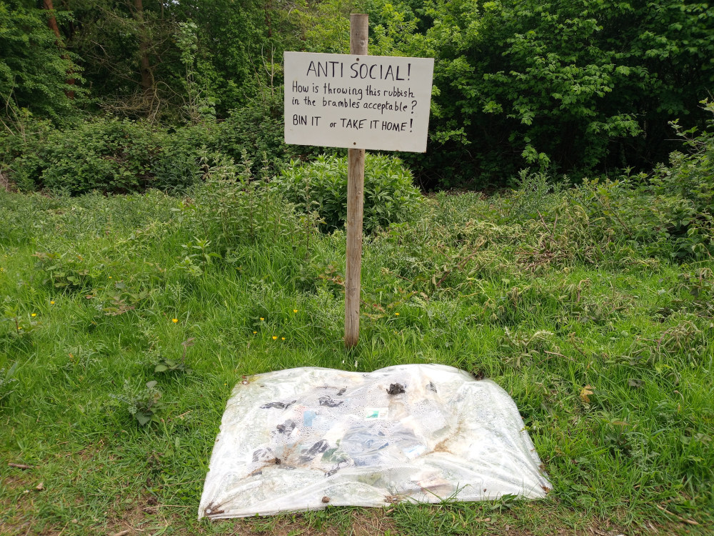 Weathered, off-white sign staked into the ground. Handwritten in black marker, the sign reads ANTI SOCIAL! How is throwing this rubbish in the brambles acceptable? BIN IT or TAKE IT HOME! Directly below the sign, a large, translucent plastic sheet lies on the ground. The sheet is irregularly shaped, rumpled, and contains a scattering of refuse—bits of plastic, paper, and possibly organic waste. This refuse is subtly visible through the plastic's translucency. The sign and the plastic sheet are the main objects highlighting the message of littering and its consequences. A simple wooden post supports the sign, driven into the earth.