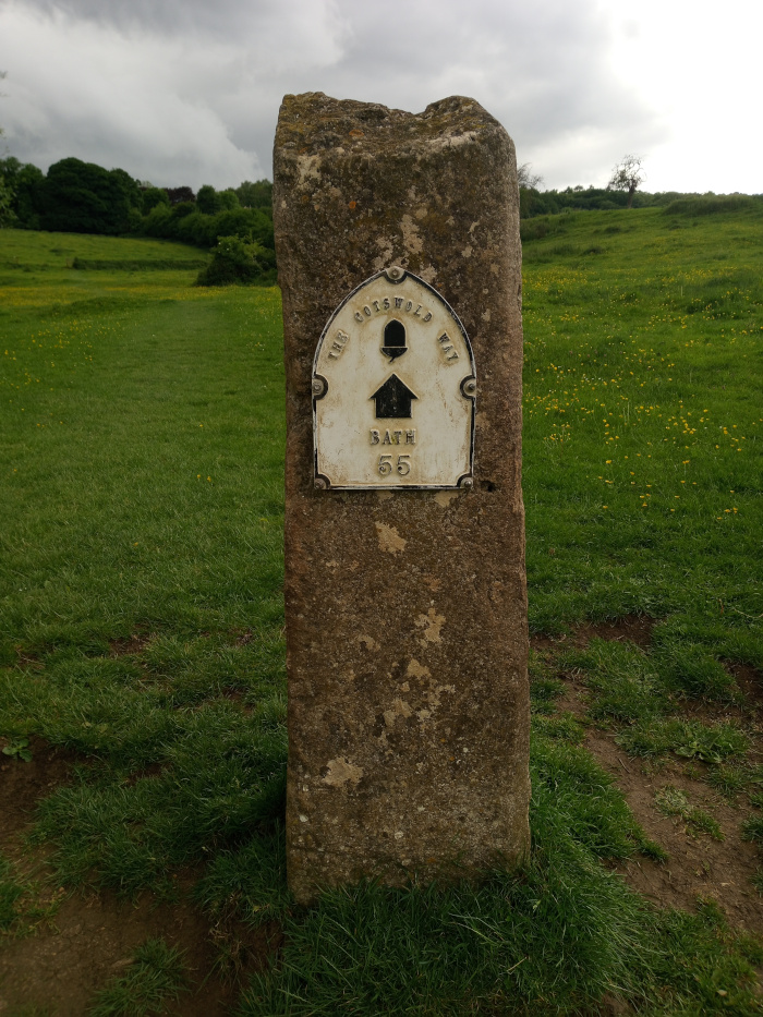 Marker is the focal point, occupying a significant portion of the frame. Its stone is a mottled, earthy brown, heavily textured with lichen or discolouration, suggesting age and exposure to the elements. The top is somewhat irregular, showing signs of natural erosion. A white enamelled plaque is affixed to the marker's face, bearing the inscription THE COTSWOLD WAY in a slightly arched semi-circular pattern at the top, flanked by decorative elements. Below, a stylised house icon points downwards, followed by the word BATH and the number 55, indicating distance or a waypoint. The plaque's white is faded, with hints of aged yellowing. The metal edging shows signs of rust or wear.