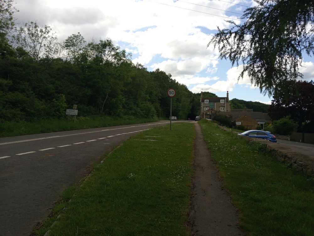 A quiet country road scene. The main focus is a paved road stretching from the foreground to the middle ground, where it curves gently to the right. White dashed lines delineate the road's center. A grassy verge, dotted with small white wildflowers, runs alongside the road on both sides. A narrow, unpaved footpath or bridleway meanders parallel to the road, also moving from the foreground towards the middle ground, seemingly leading to a pub.
