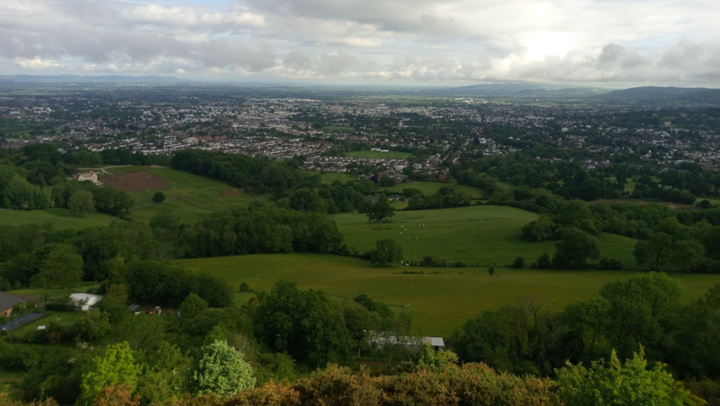 Panoramic view from an elevated position, possibly a hilltop. It shows a sprawling landscape with rolling green hills dotted with trees and patches of farmland. In the distance, a large city or town is visible, extending to the horizon. The sky is mostly cloudy with varying degrees of cloud cover. The overall impression is one of a serene, expansive countryside scene contrasting with the urban sprawl in the background.