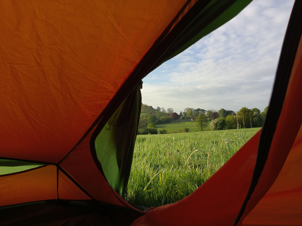 Interior of a brightly coloured, orange and green dome tent. The tent fabric is slightly wrinkled and shows the texture of nylon or a similar material. A section of the tent's zip is visible, suggesting the tent is partially opened.  No people or other objects are inside the tent. The background seen through the opened section of the tent is a picturesque pastoral scene. A field of vibrant, spring-green grass stretches from the bottom edge of the frame to a line of low-lying hills in the distance. On these hills, we see a cluster of quaint buildings — possibly a small village or farmstead — with a scattering of trees, creating a naturalistic, bucolic landscape. The sky is a soft, pale blue, lightly clouded, with hints of white and grey, suggesting a calm, possibly early morning or late afternoon light.