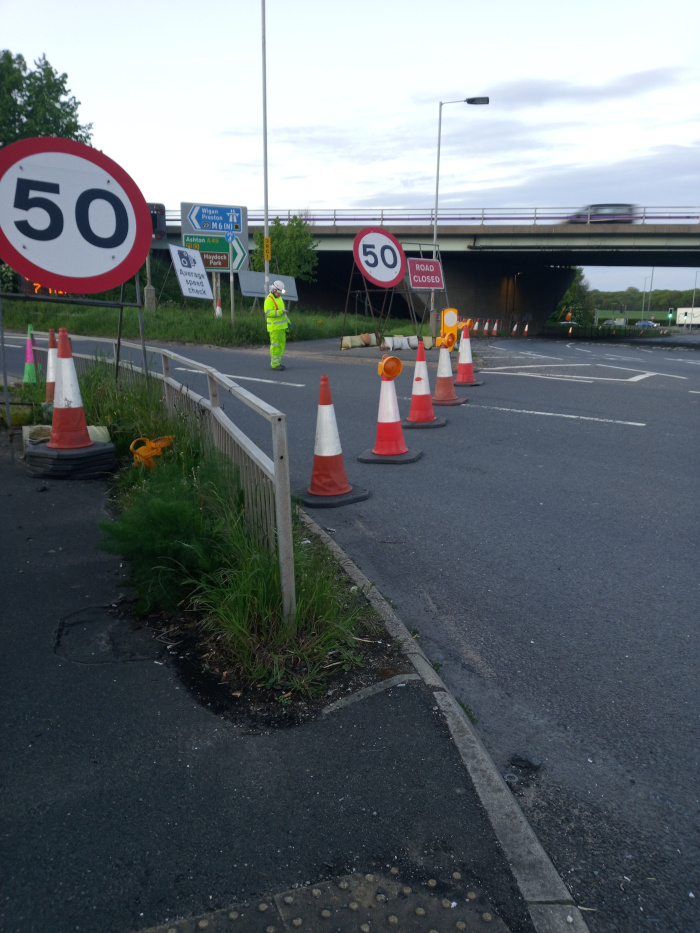 Lone worker in a bright neon-yellow high-visibility jacket and helmet, standing calmly near a row of traffic cones. They appear to be observing or directing traffic. Their body posture suggests attentiveness and a state of readiness. The worker is positioned slightly off-center, creating a dynamic rather than symmetrical composition. Several traffic cones, varying in heights and shades of orange and red, extend from the worker's position towards the right edge of the frame, forming a gently curving line. Two large circular signs reading 50 indicating speed limits are prominently featured; one is to the left, in the foreground, another, farther back, just before the road closure signs. Several additional smaller road signs are present, displaying information regarding directions, local areas, and average speed checks, suggesting a road junction. These signs are mounted on metal posts, varying in height and positioned on either side of the road.