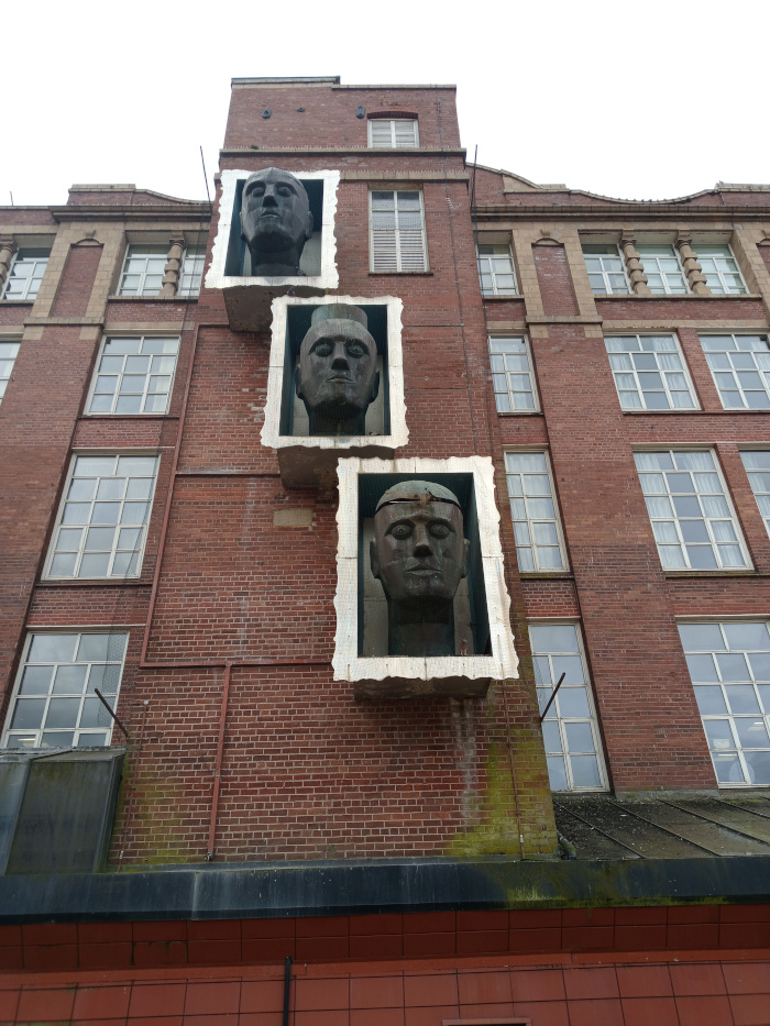 Three bronze-toned, sculpted heads, each encased within a white, roughly hewn, rectangular frame that's recessed into the brick facade of a building. The frames appear slightly distressed, adding texture. The heads are positioned vertically, one above the other.